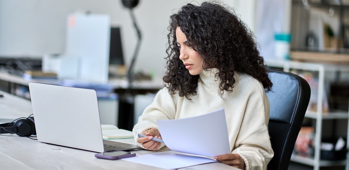 Young latin business woman holding documents working with laptop.